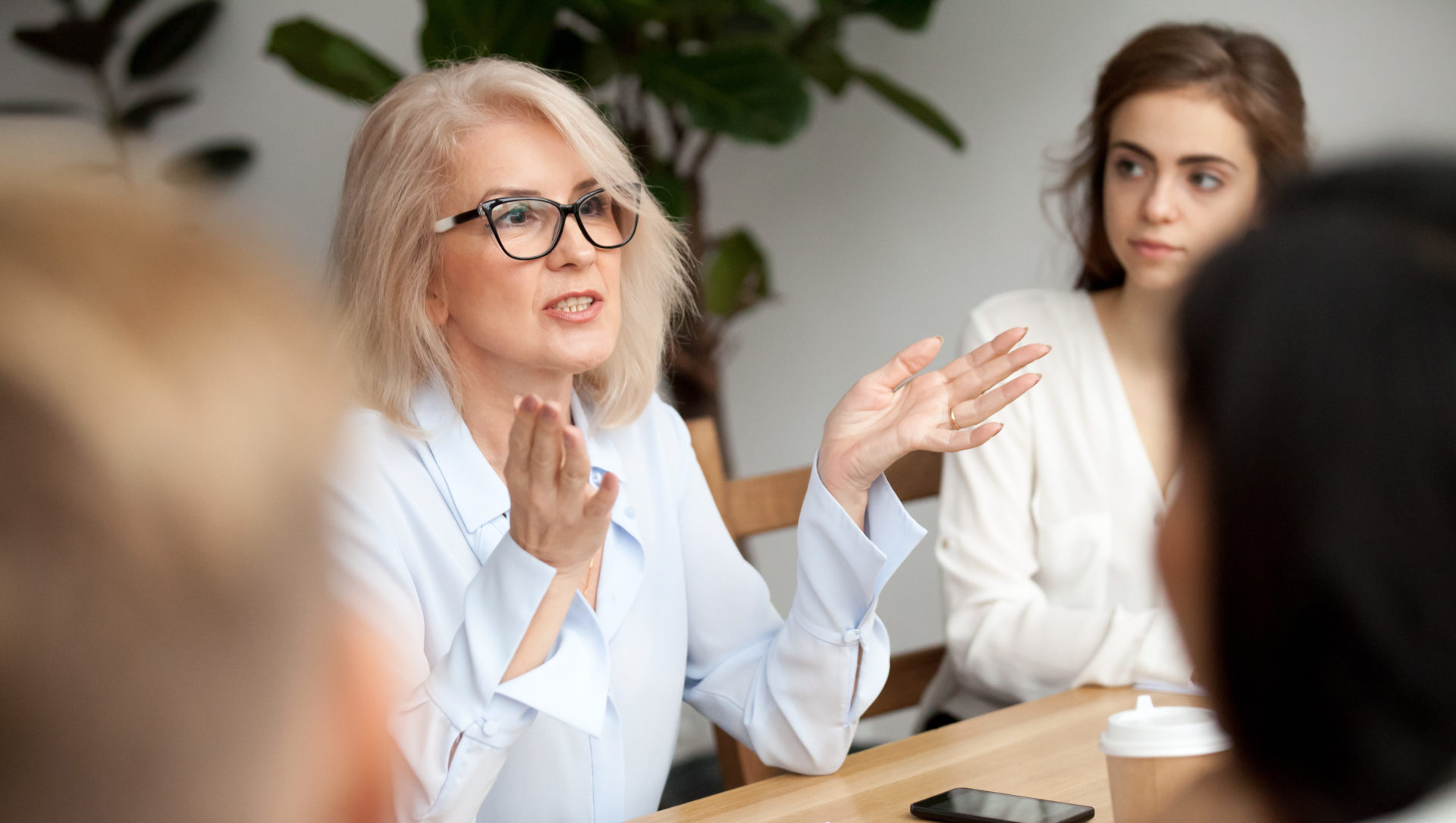 Attractive aged businesswoman, teacher or mentor coach speaking to young people, senior woman in glasses teaching audience at training seminar, female business leader speaker talking at meeting