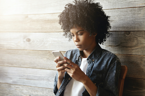 Beautiful young African hipster girl with curly hair wearing trendy clothing holding mobile phone, reading sms from her boyfriend, having serious and worried facial expression. People and technology