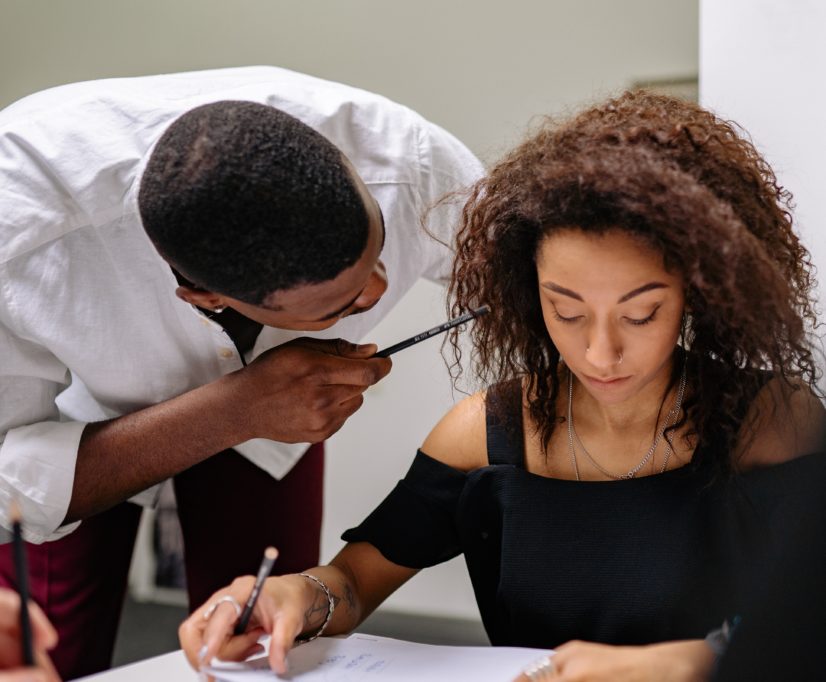 A woman sitting at a desk and a man bent over her looking like he's agressively saying something close to her face