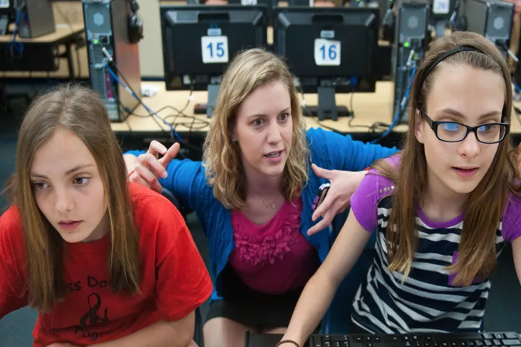 two female students being helped at their computers by a teacher in class
