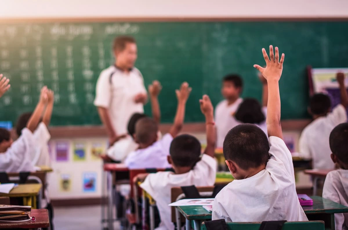 Students raising hands in classroom with teacher.