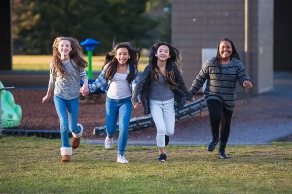 Four smiling diverse girls running and holding hands in a park, illustrating friendship and social connection.