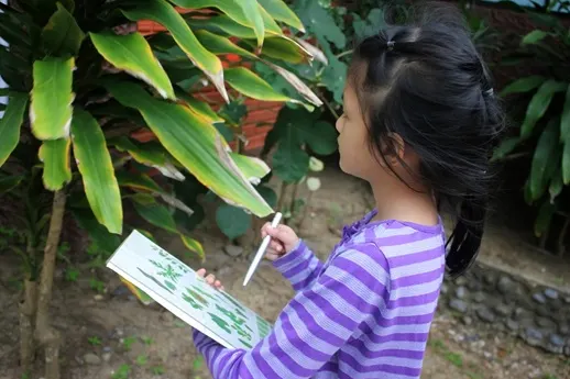 A young student holds a notebook and a pen, appearing to draw or observe plants outdoors.