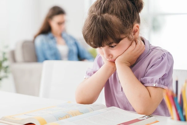 A young student looks bored or sad while doing homework at a desk, with an adult out of focus in the background.