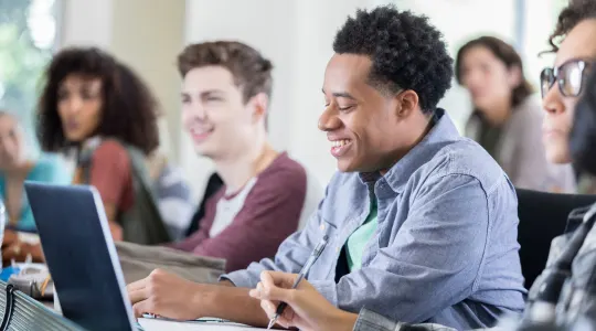 A diverse group of young adults sitting in a classroom, with one male student smiling while looking at a laptop.