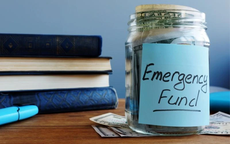 A clear jar with a label that says "Emergency Fund" filled with money, sitting on a desk next to a stack of books and scattered cash.