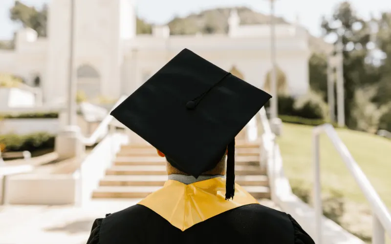 A graduating student in a cap and gown walking toward a building, seen from the back.