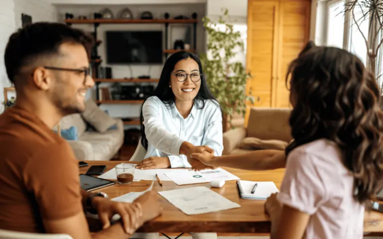 A financial professional in glasses smiles and shakes hands with a customer across a table, representing a personal financial consultation or closing a deal.