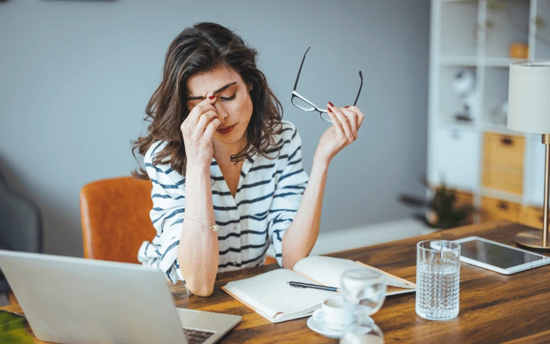 A stressed woman holds her glasses and rubs her eyes while sitting at a desk with a laptop and open notebook.