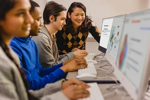 A teacher helping three diverse students working on computers with data and graphs.