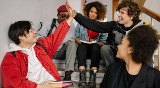 A diverse group of young students sitting on stairs, smiling and high-fiving.