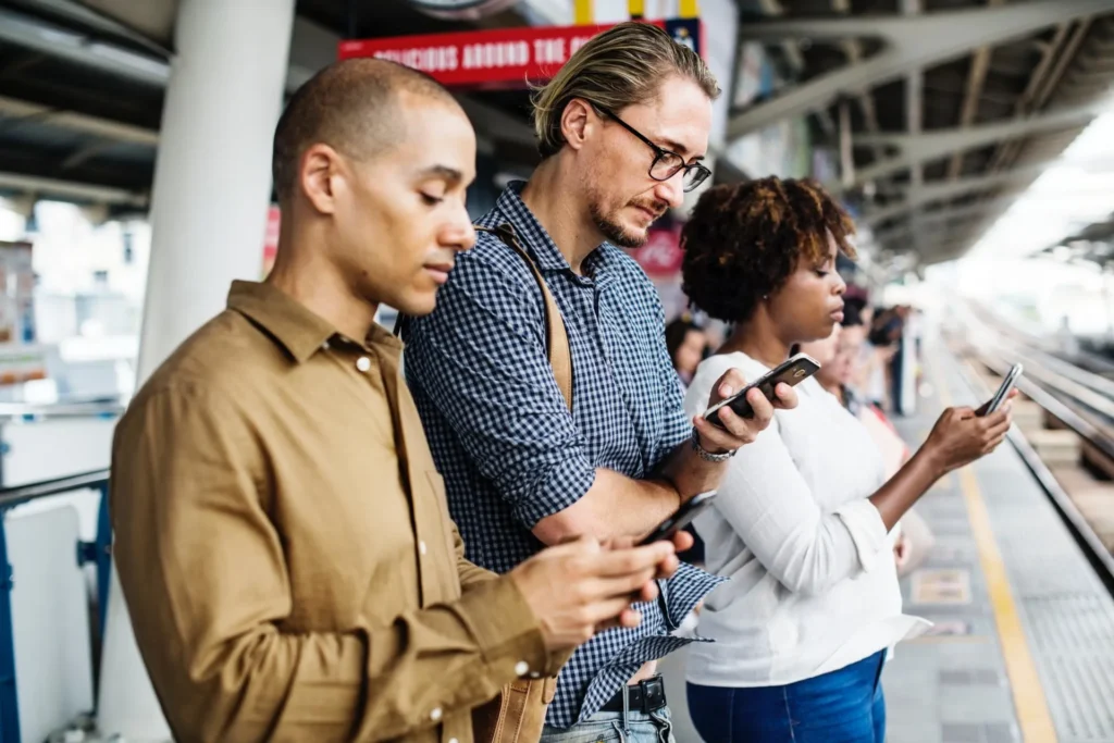 Three millennials standing on a subway platform, each focused on their smartphones, symbolizing digital habits